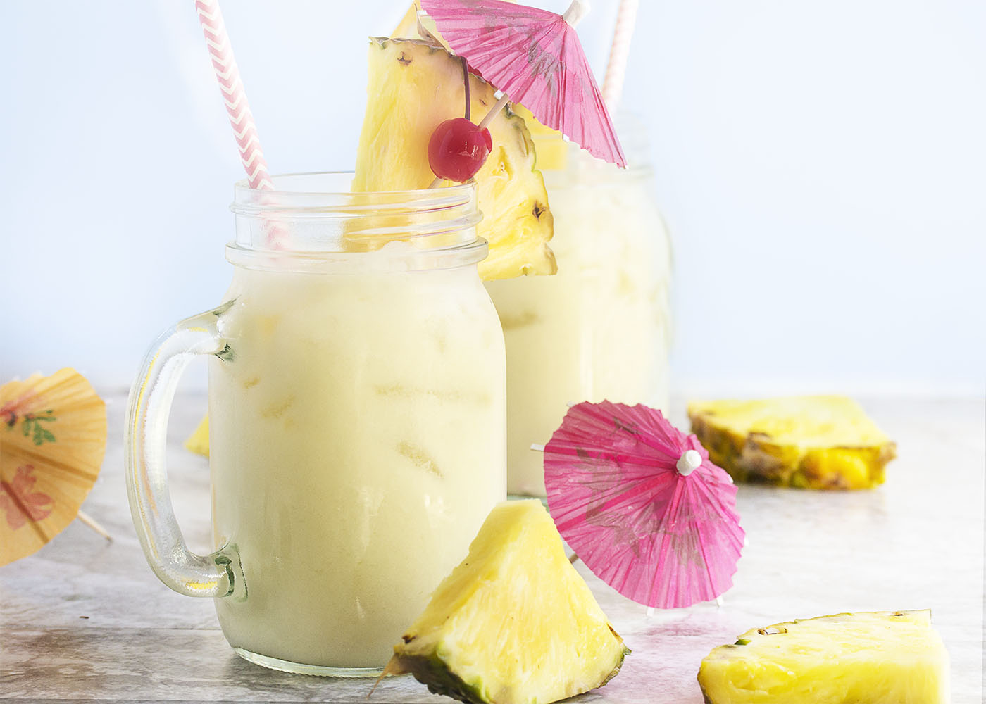 View of two pina coladas in mason jar glasses with straws, umbrellsas, and pineapple slices.