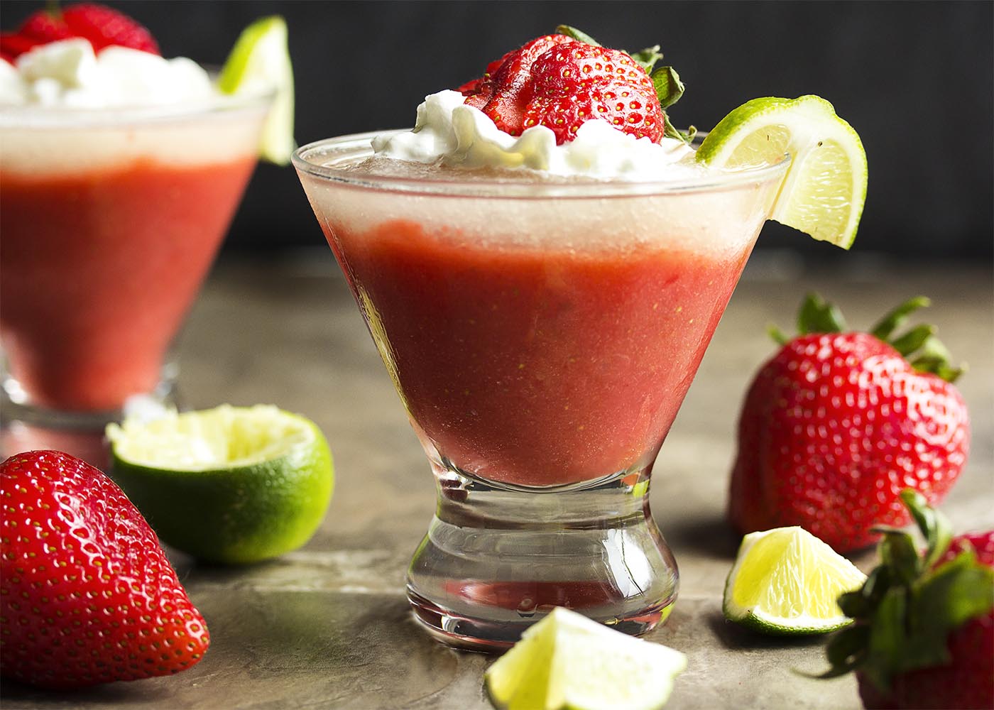 Two strawberry daiquiri cocktails each layered with lime and garnished with strawberries and whipped cream.