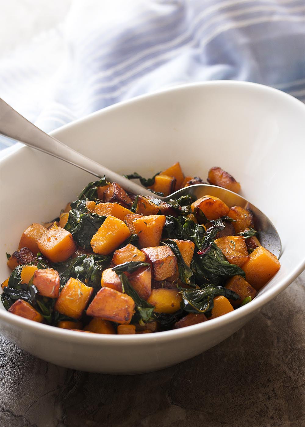 Side view of sauteed butternut squash cubes and Swiss chard in a white serving bowl with a serving spoon