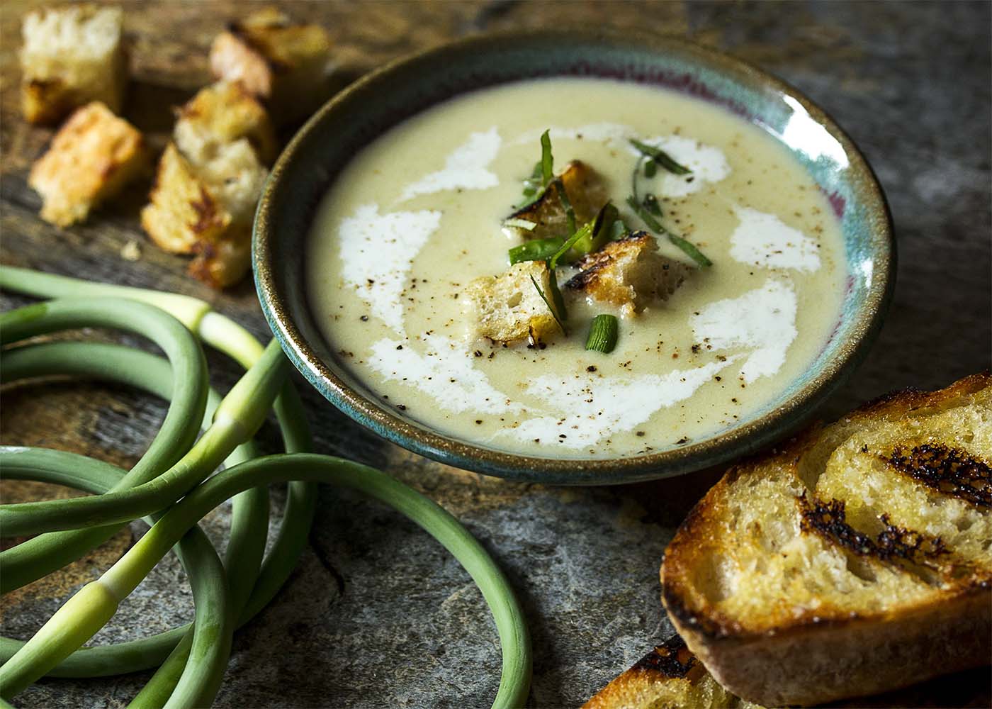 A bowl of cold garlic scape soup topped with garnishes. Grilled bread and garlic scapes scattered about.