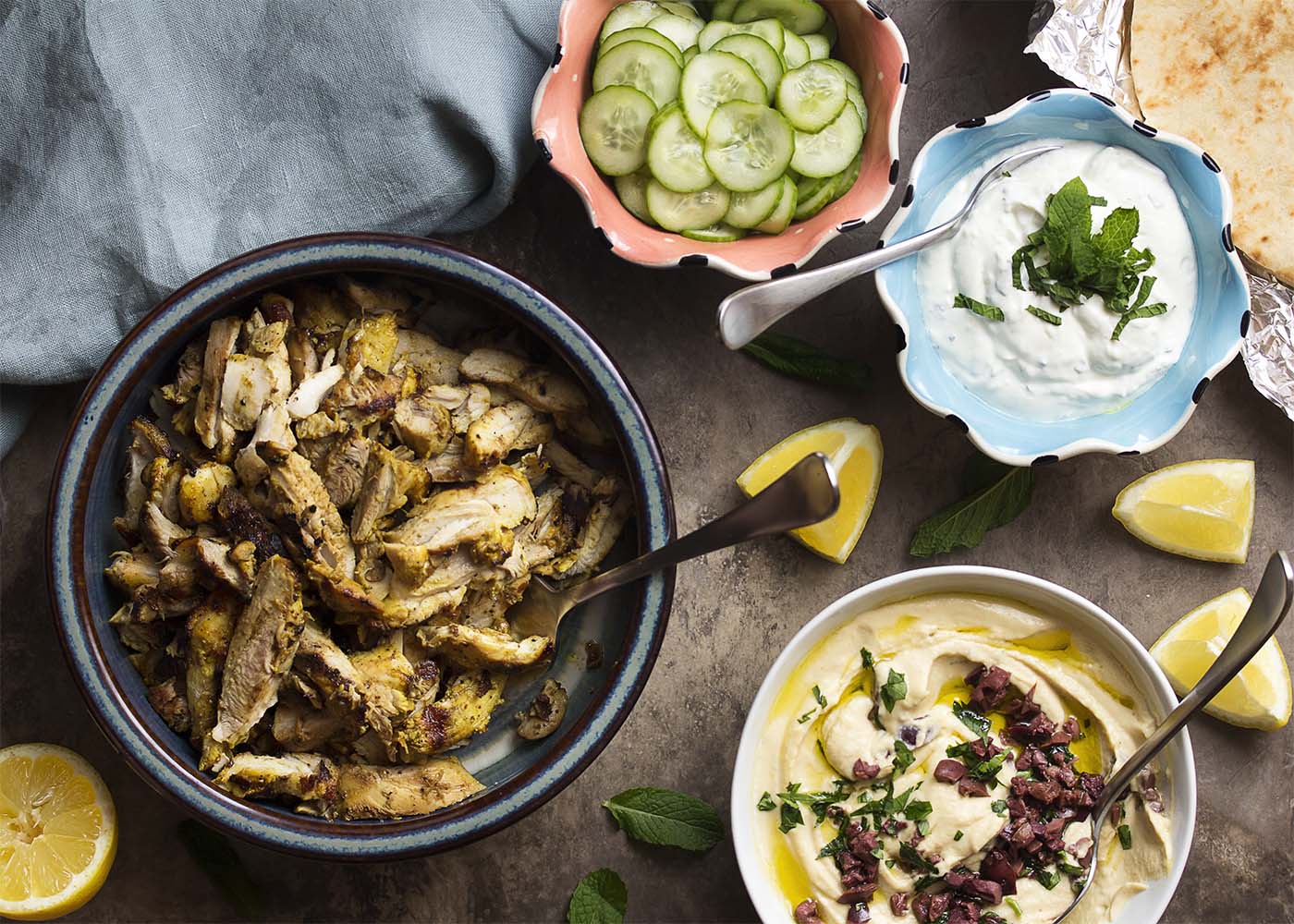 Top view of a table covered in platters of chicken shawarma, pickled cucumbers, yogurt sauce, hummus, and pitas.