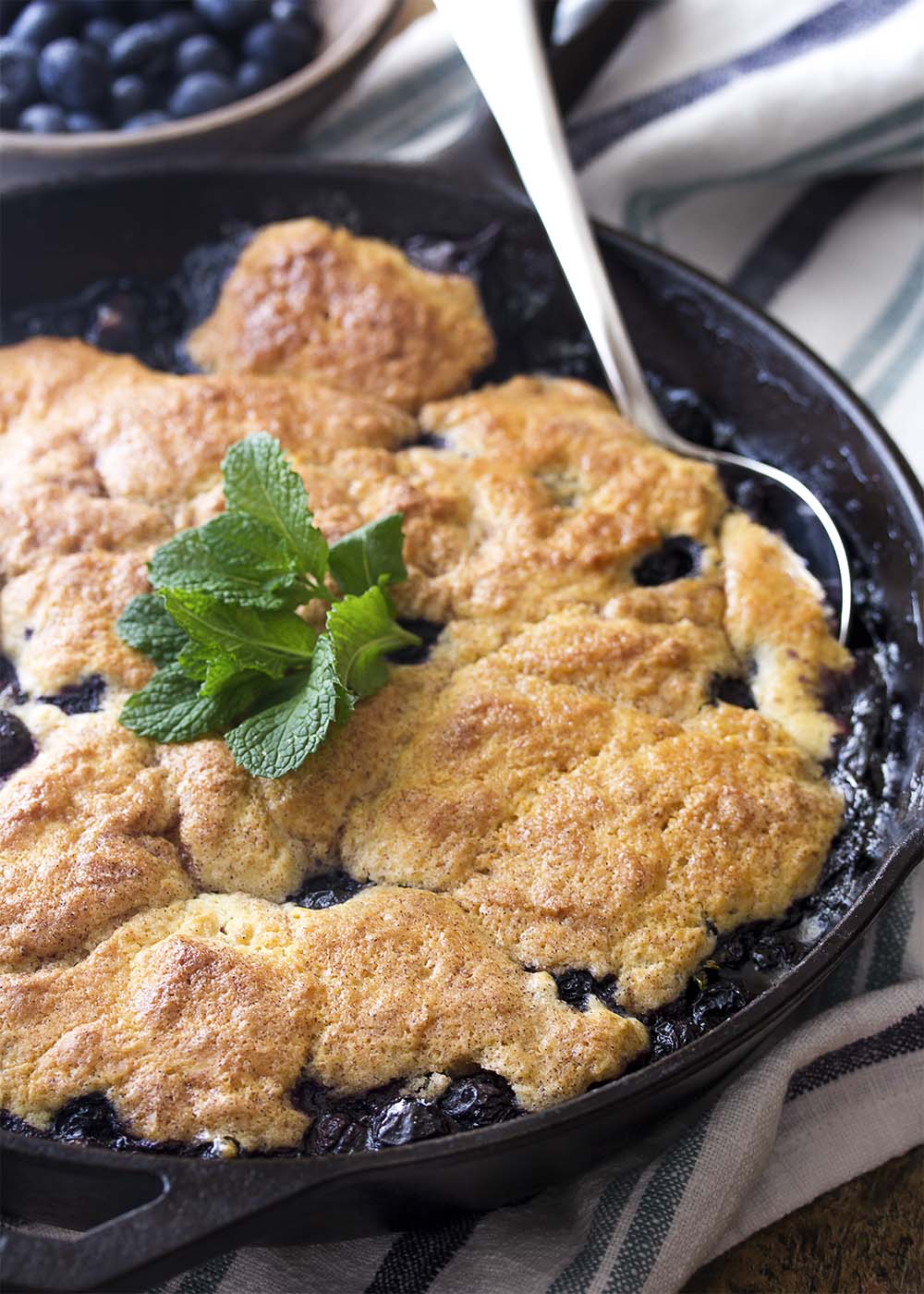 A cast iron skillet full of baked blueberry cobbler. A serving spoon in the skillet ready to scoop some out.