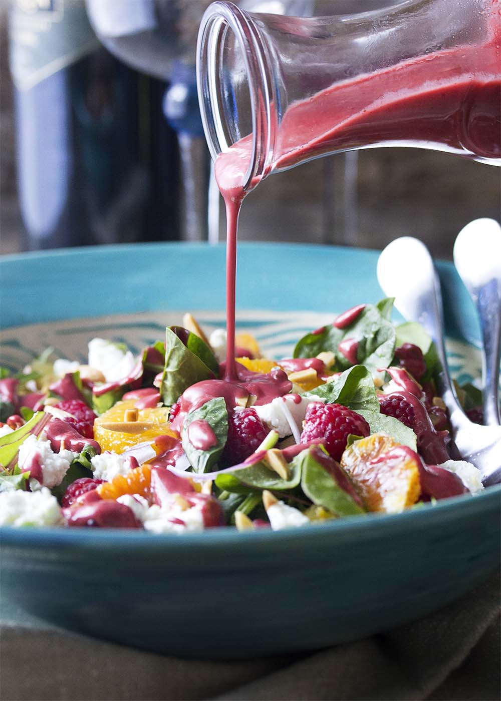 Thick, deep pink raspberry vinaigrette being poured over a large serving bowl full of mixed greens and raspberry salad.