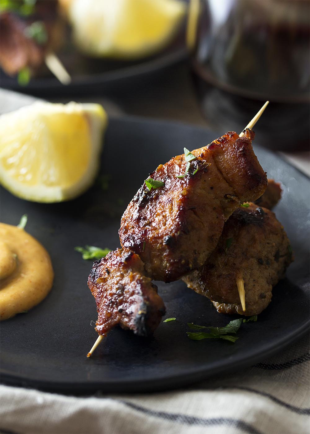 Close up of two skewers of pinchos morunos on a small black plate. Wedge of lemon and dollop of aioli in the background.