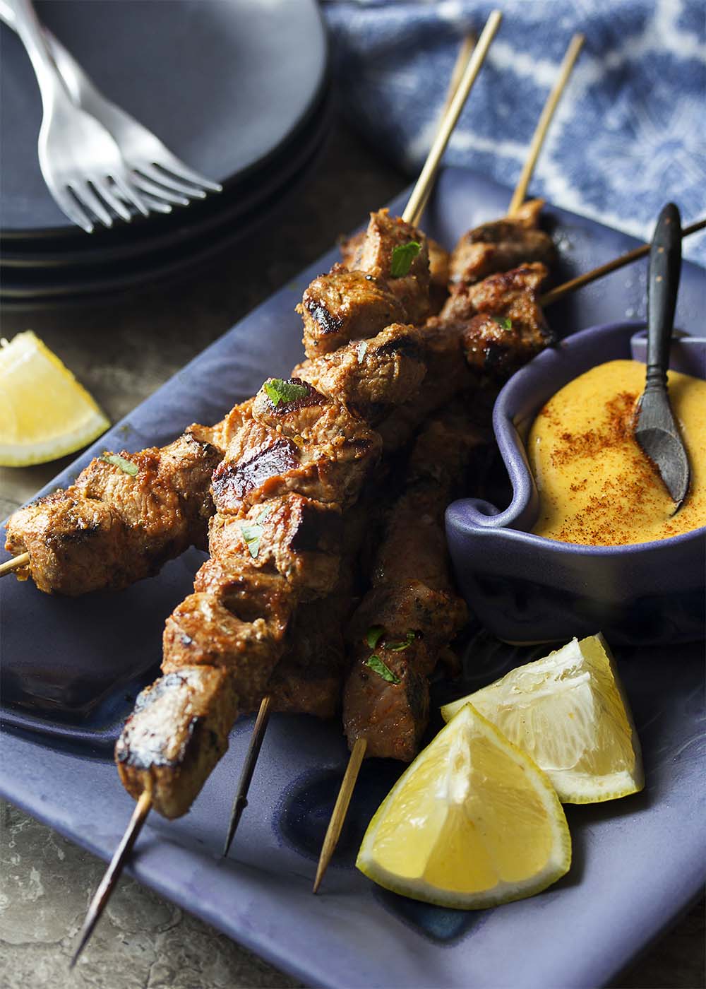 Long skewers of pinchos morunos on a blue serving platter with wedges of lemon and a bowl of aioli.