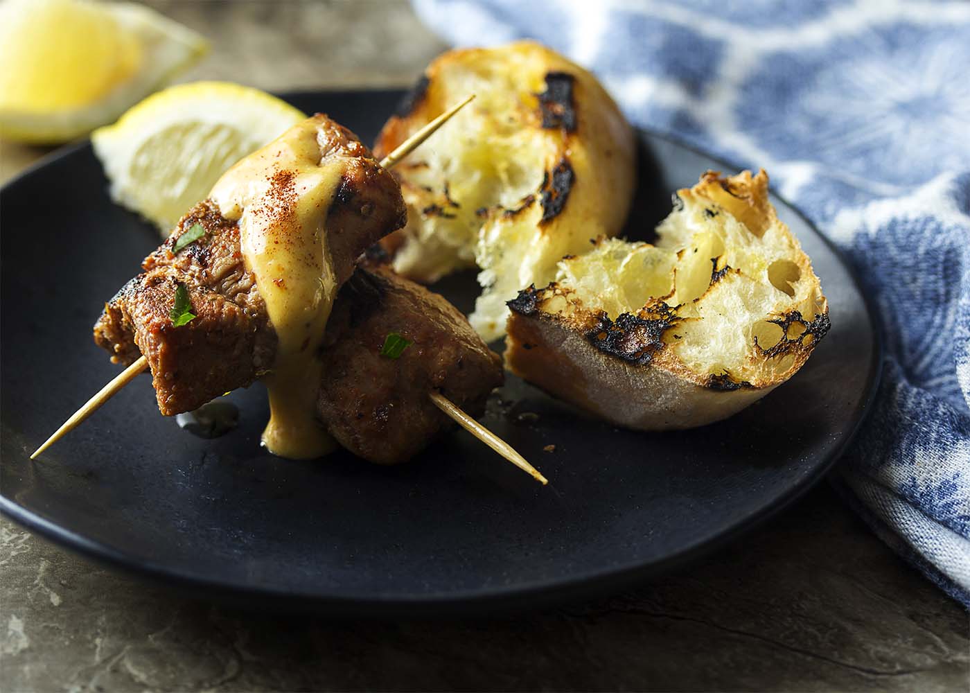 Two skewers of pinchos morunos with aioli dripping down them and grilled bread to mop up the sauce.