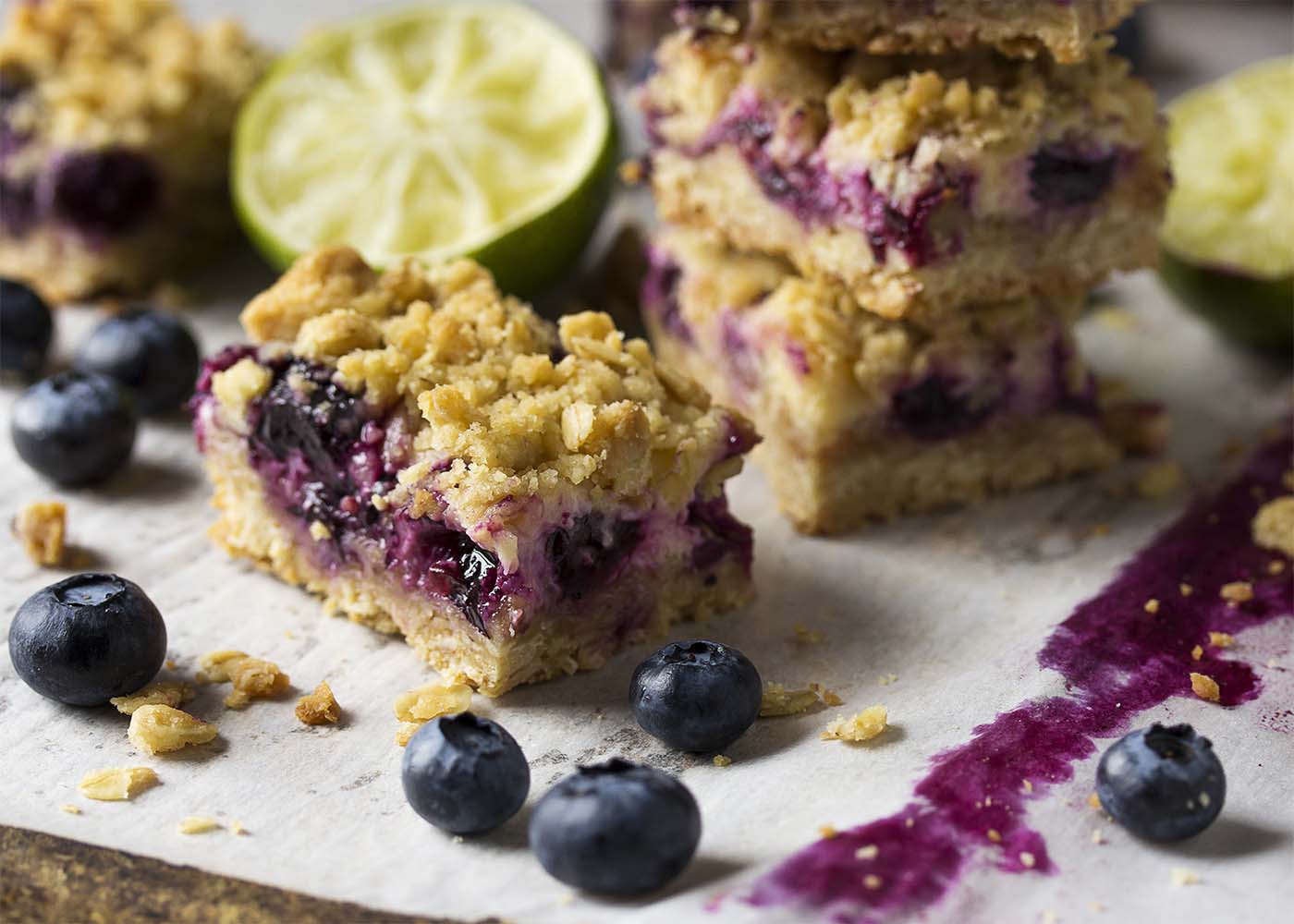 One blueberry lime oatmeal bar on parchment paper. Blueberries, crumbs, and other bars scattered about.