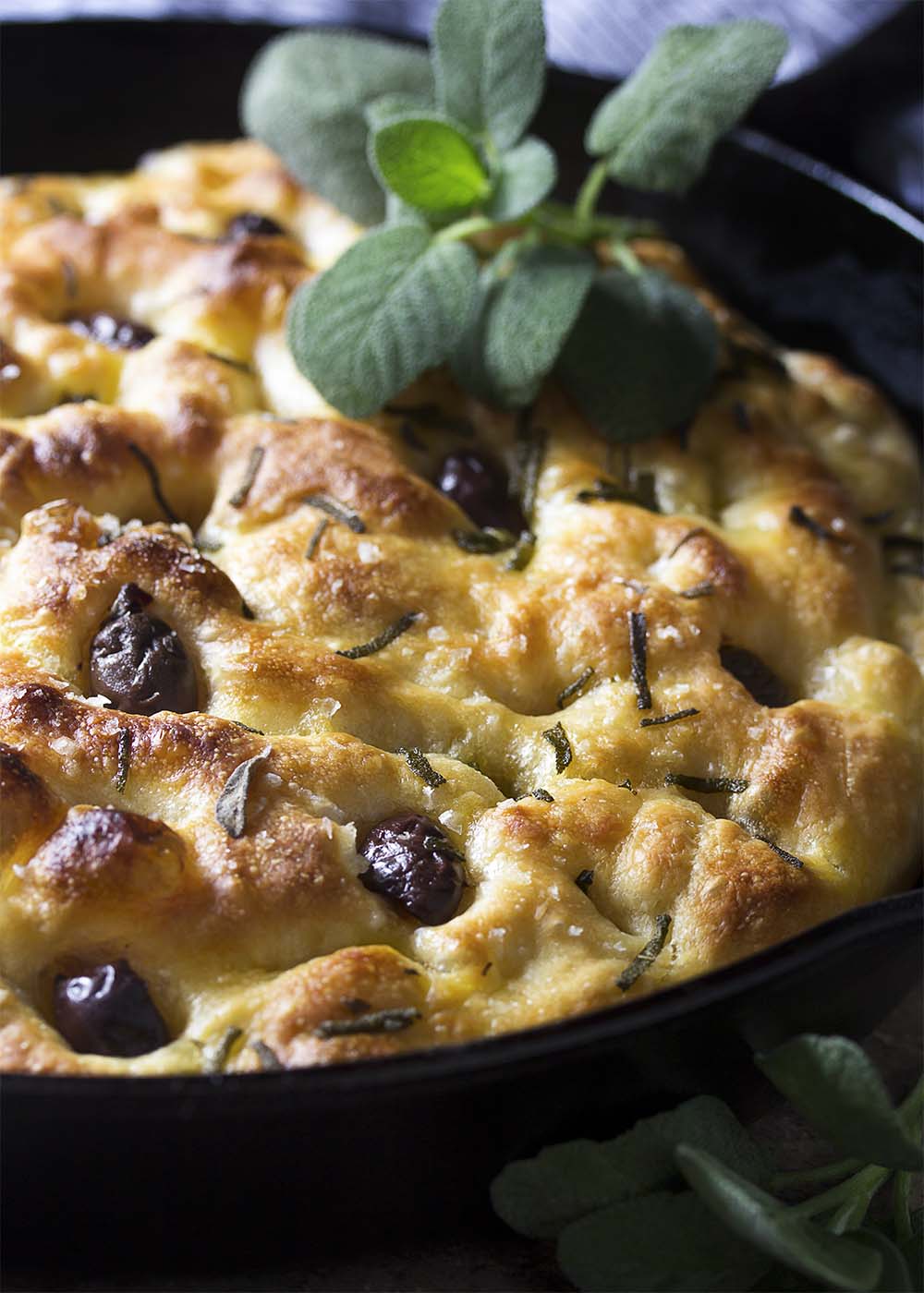 A golden brown loaf of sage and olive focaccia in a cast iron skillet.