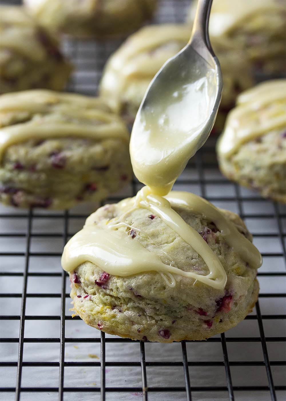 A spoon drizzling white chocolate onto a cranberry scone on a cooling rack.