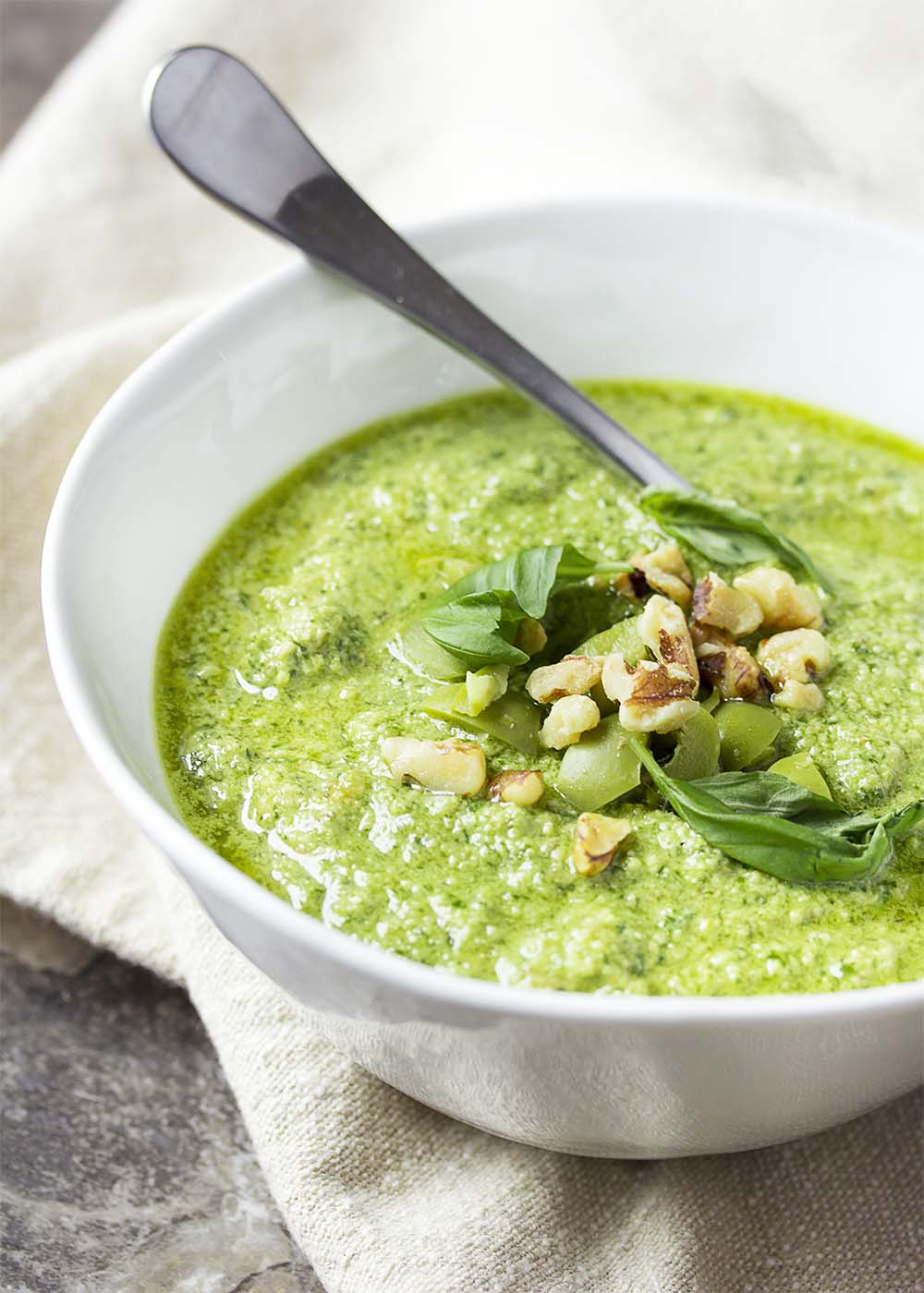 White bowl of bright green pesto with a spoon. Sauce topped with basil and walnuts.
