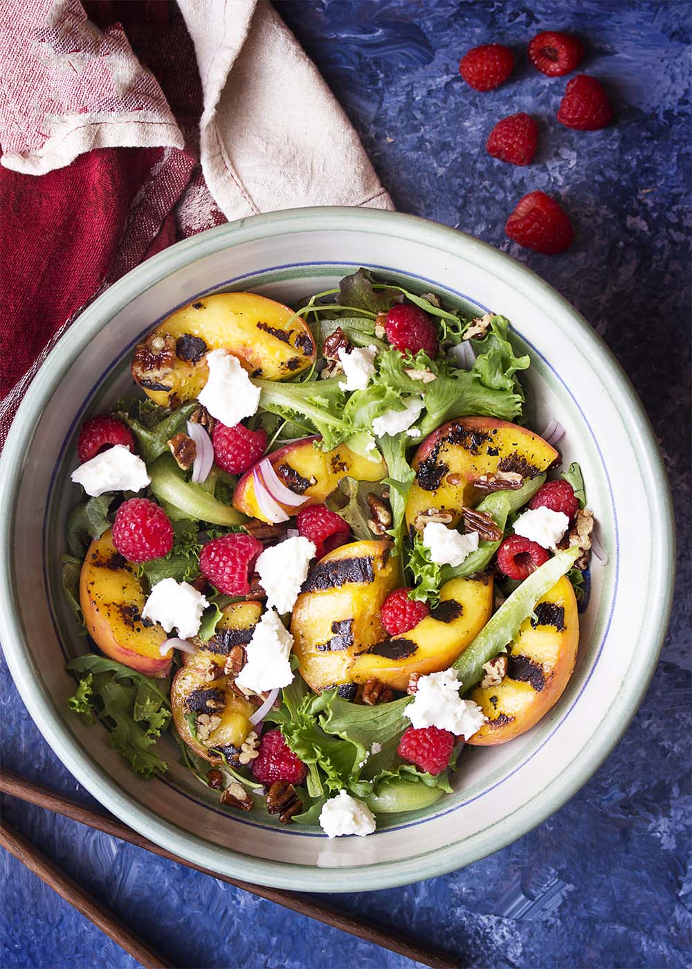 A serving bowl of salad with peaches, raspberries, goat cheese, red onions, pecans, and mixed greens. Serving tongs and a cloth napkin on the table nearby.