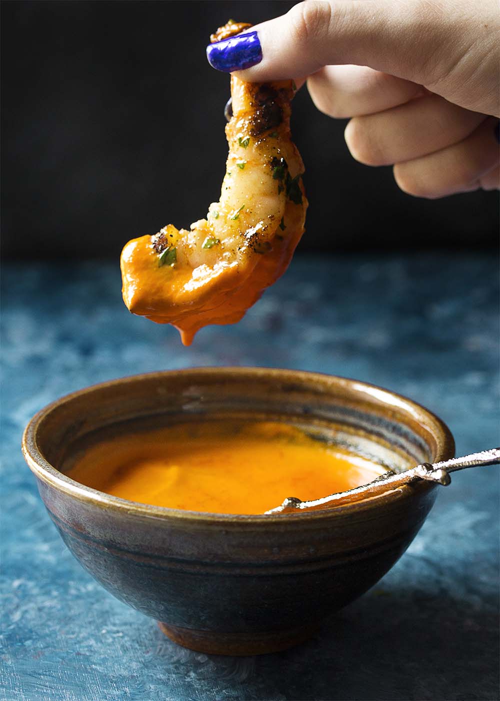 An hand holding up a grilled shrimp in the air just above a bowl of dipping sauce. Dipping sauce is dripping off the shrimp.