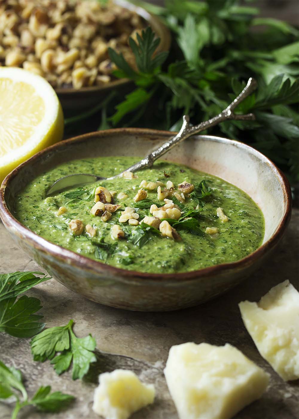 A small bowl of Italian parsley pesto topped with chopped walnuts and parsley. Parmesan cheese, parsley, and walnuts arranged around the bowl.