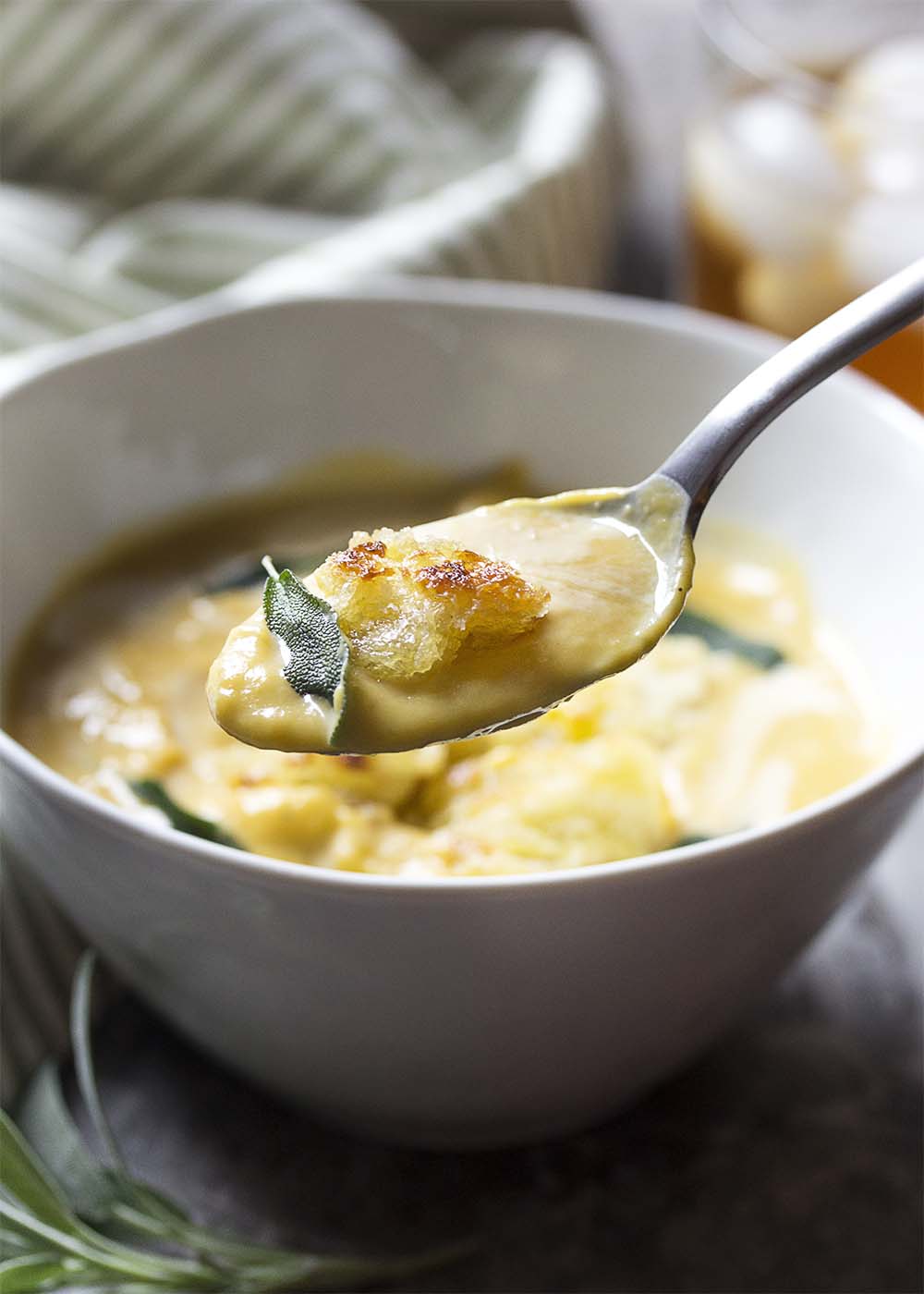 A spoon holding up some thick and creamy soup over the bowl. A crouton and a fried sage in the spoon as well.