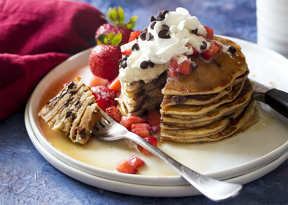 A stack of fluffy pancakes on a white plate with whipped cream, strawberries, and maple syrup. Pancakes are cut open to see the tender interior and chocolate chips.