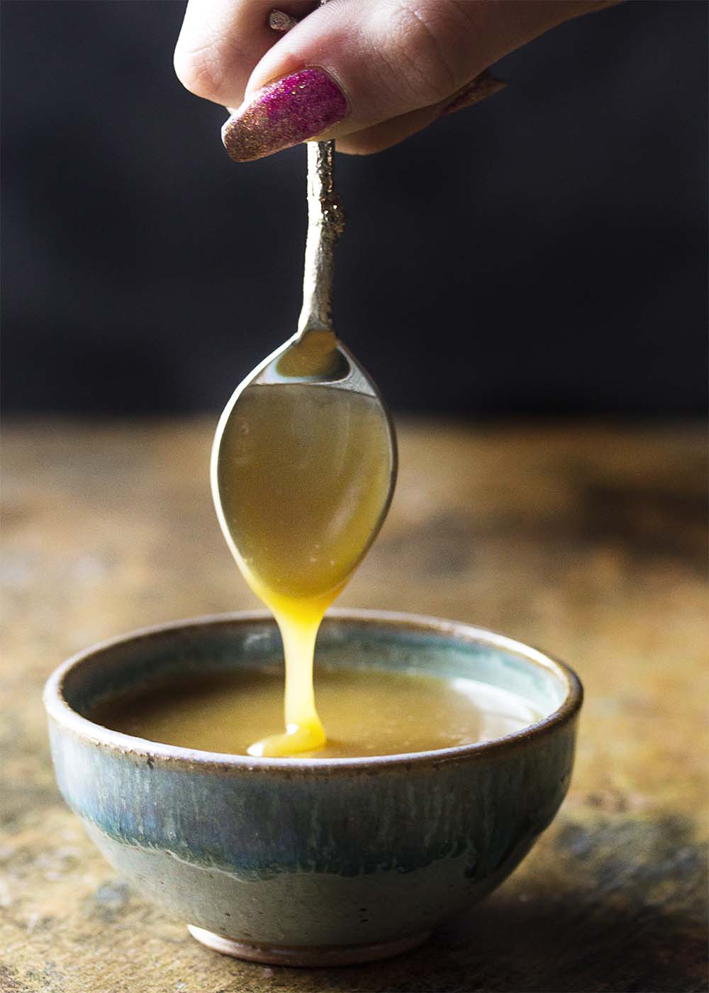 A hand holding a spoon of butterscotch sauce so that the sauce pours into a bowl.