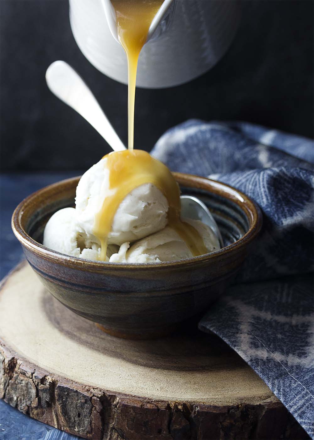 A cup pouring dessert sauce onto a bowl of vanilla ice cream.