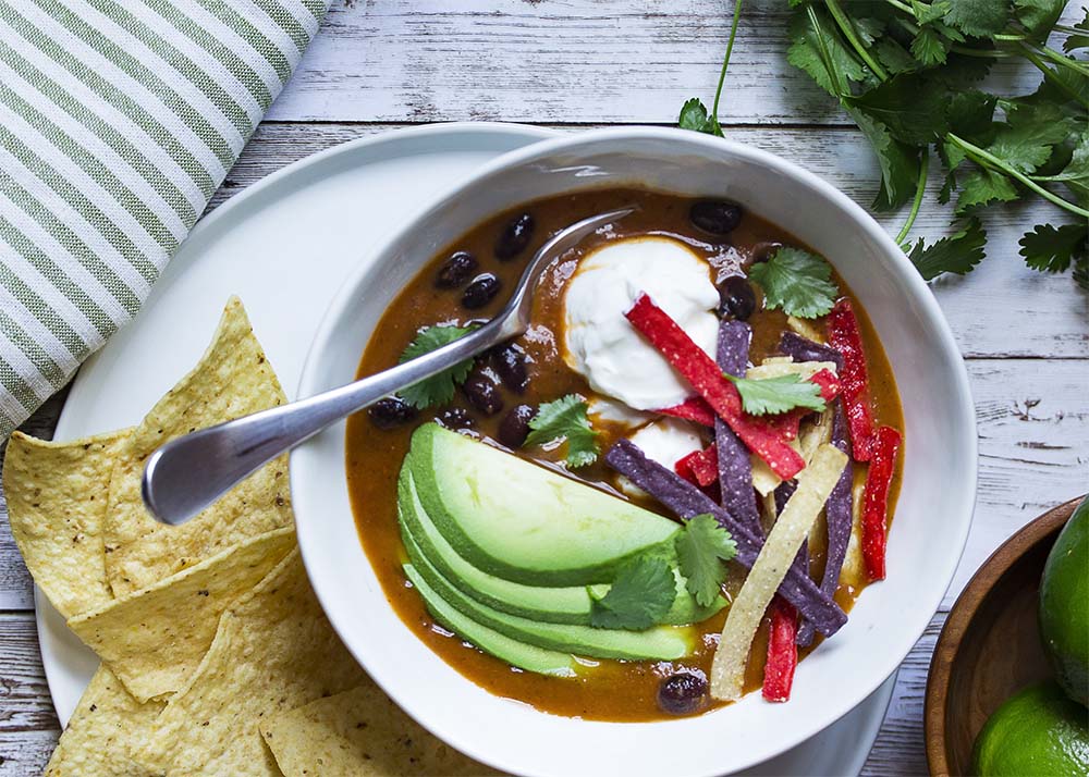 Top view of a bowl of loaded black bean soup on a plate with tortilla chips.