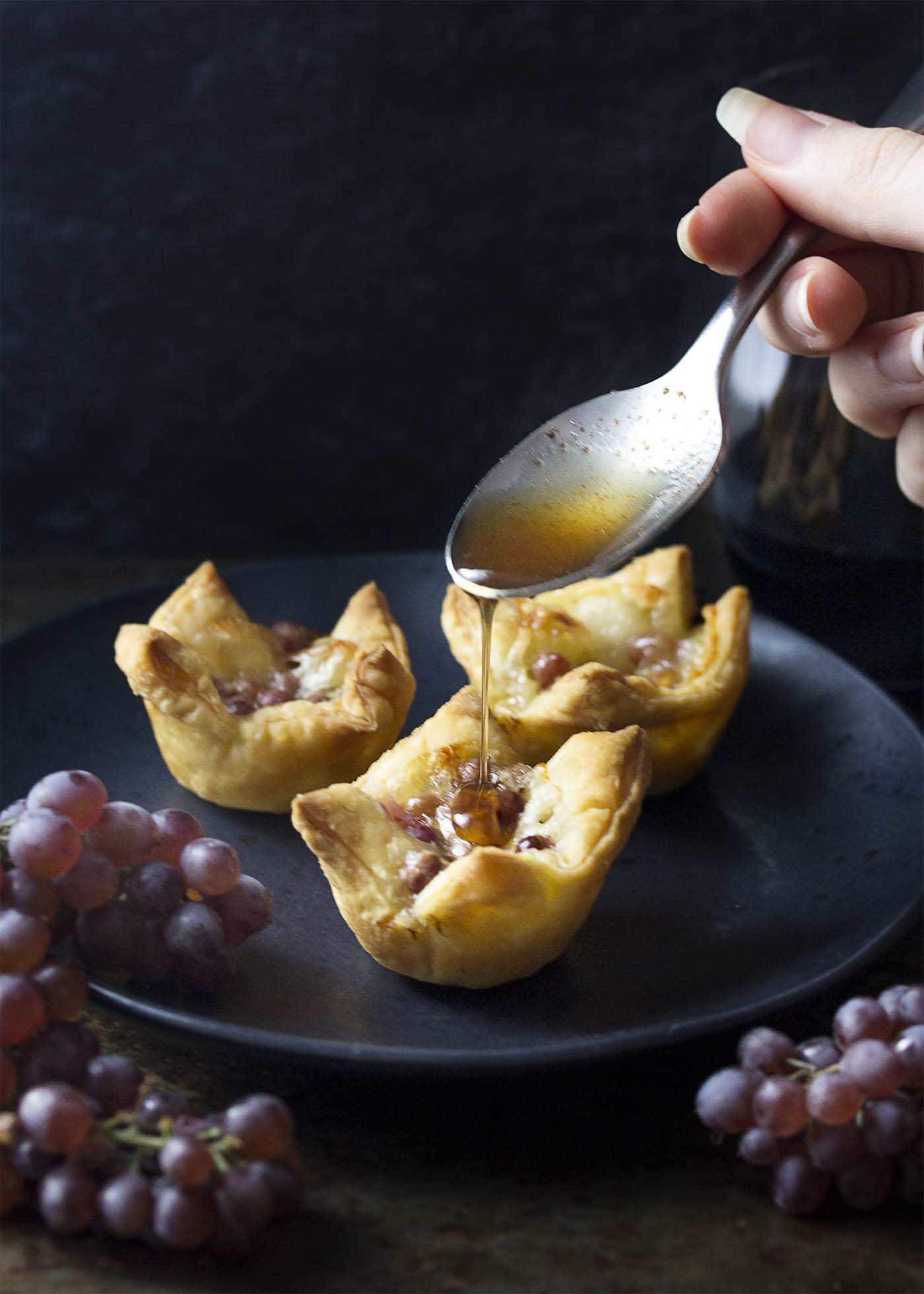 Three grape and brie bites on a black plate with spoon drizzling honey over them.