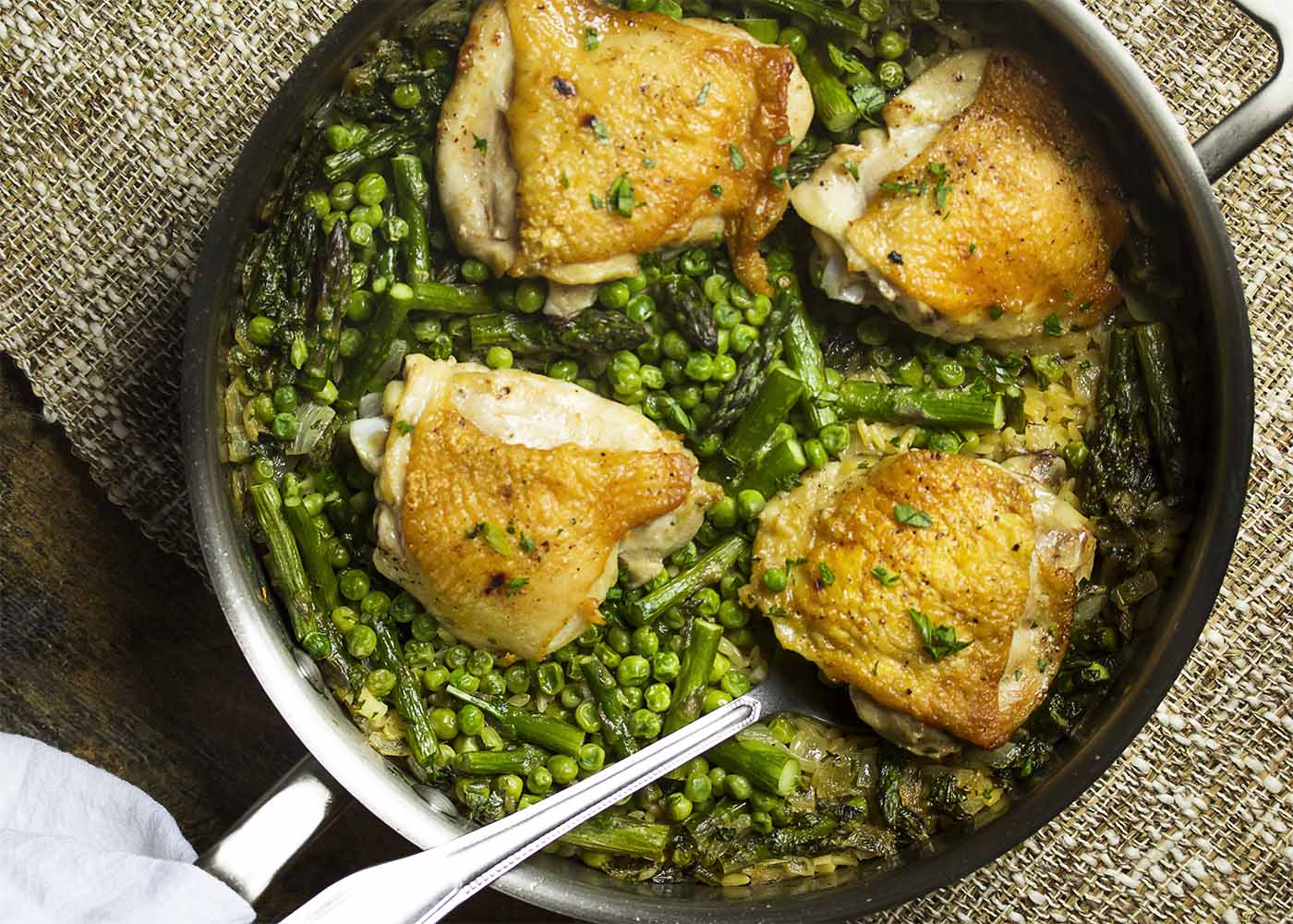Top view of a skillet filled with chicken thighs, orzo, asparagus, and peas.