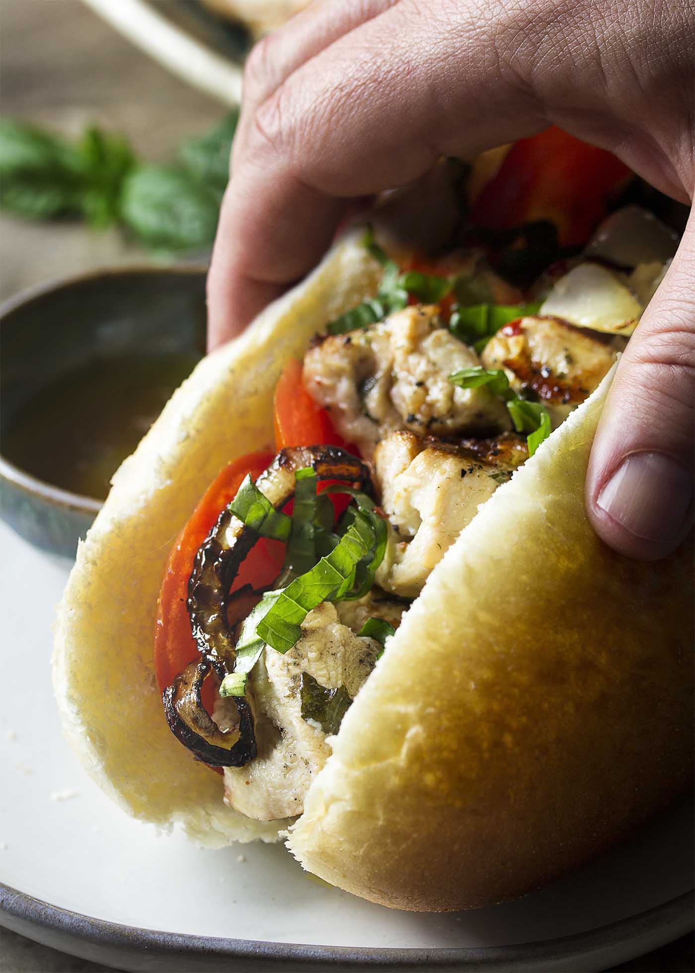 A hand lifting a bun filled with grilled chicken, onions, and tomatoes off a plate.