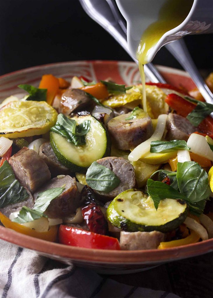 Lemon dressing pouring into a large serving bowl of grilled sausage and vegetable salad.