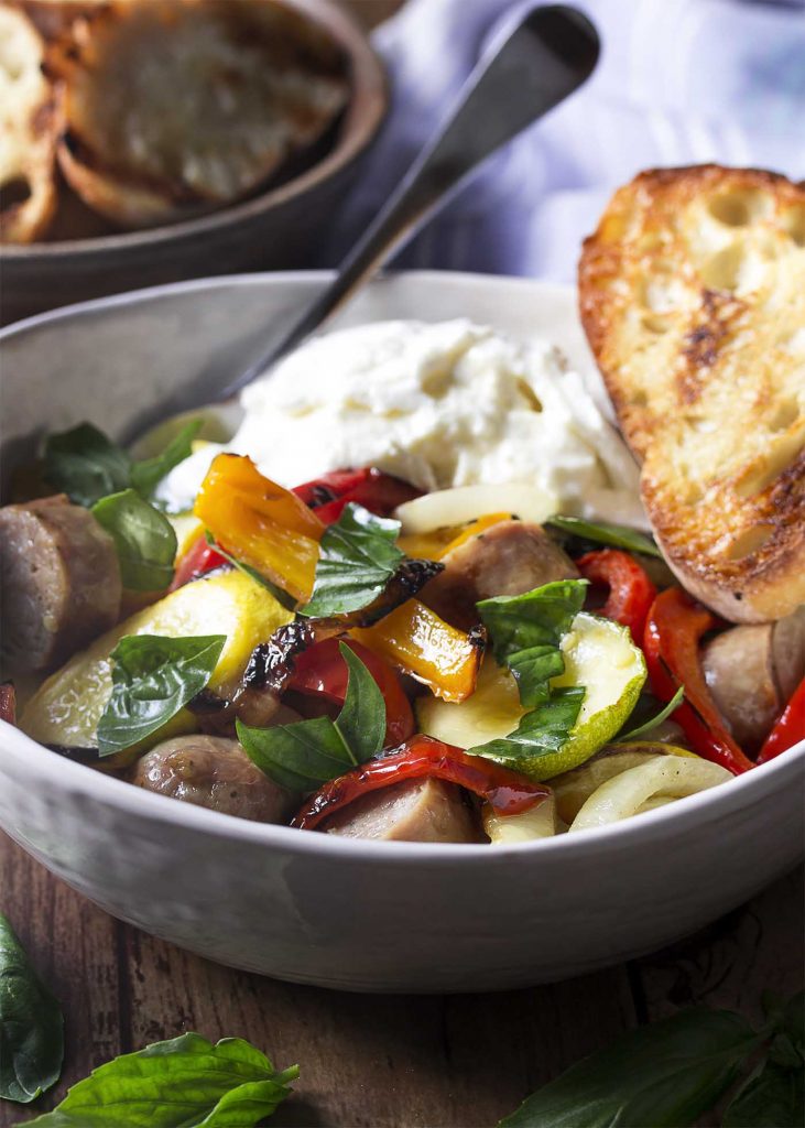 Close up of a white bowl filled with grilled sliced zucchini, peppers, and sausage topped with burrata.