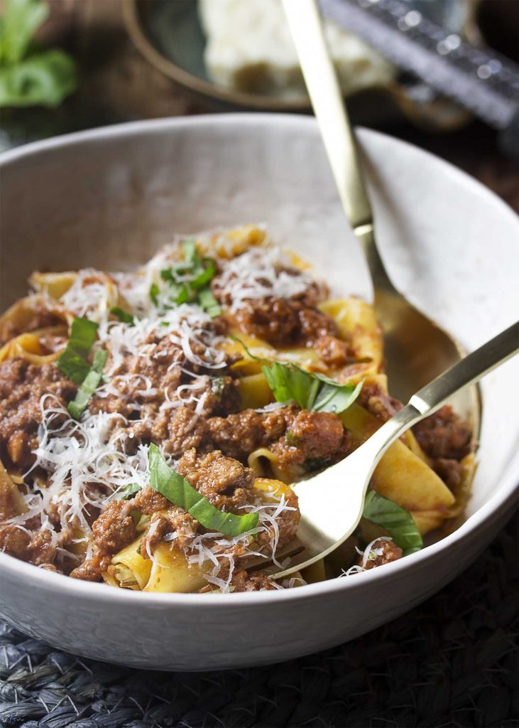 Meat sauce over pappardelle topped by shredded parmesan and basil all in a bowl.